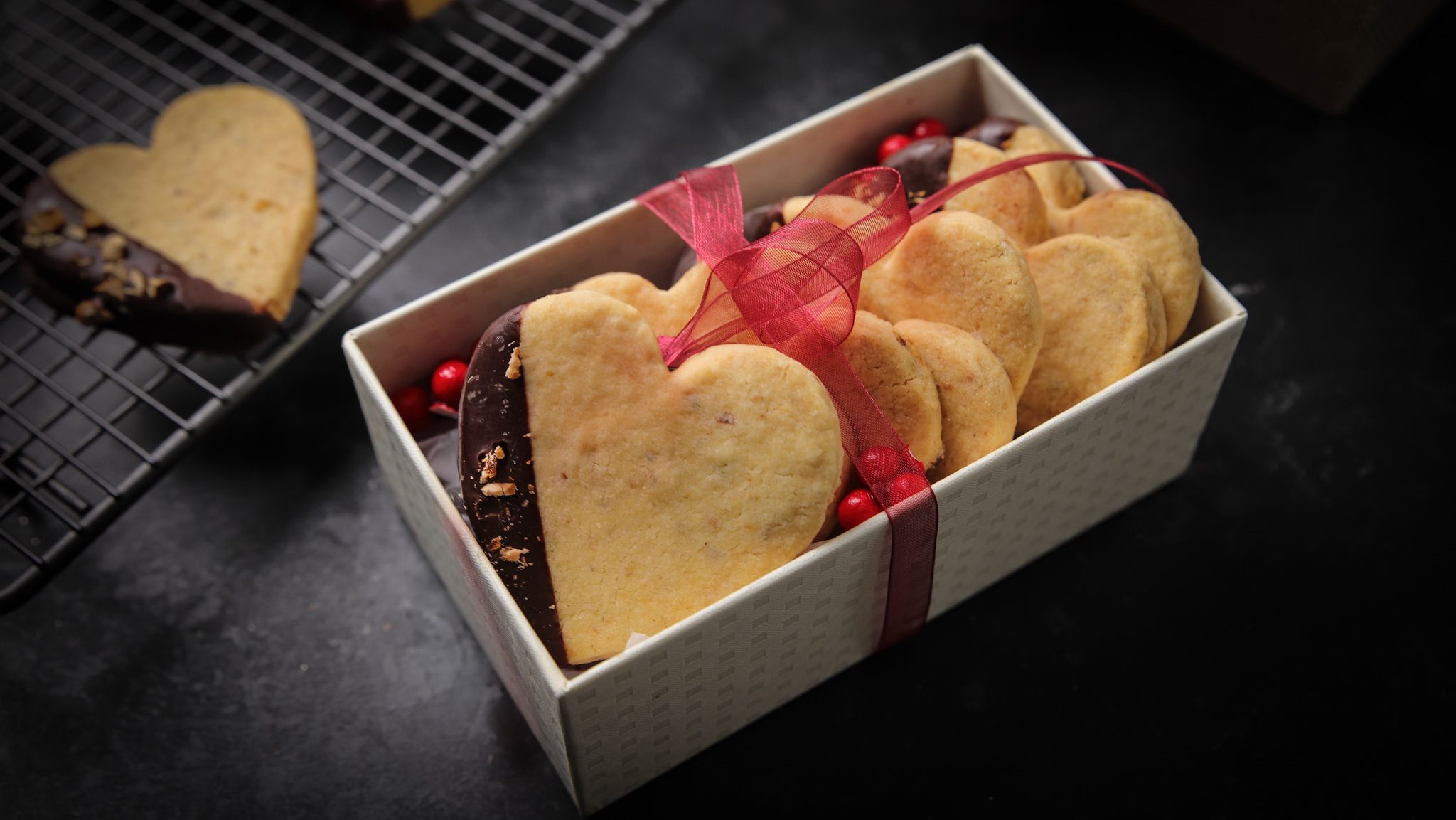 Heart shaped partly chocolate covered cookies in a gift box with a red bow. More cookies on wire rack.