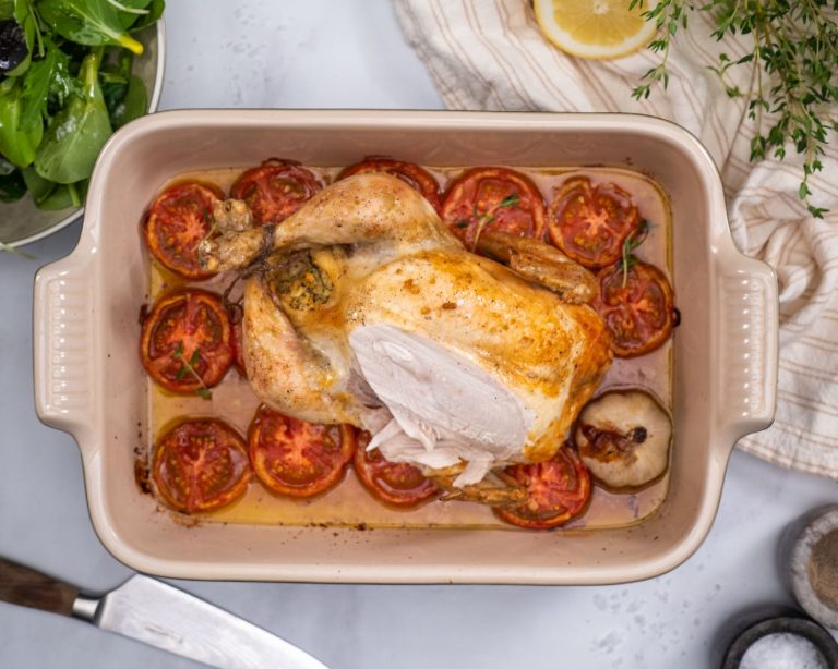 A whole roast chicken set in a deep rectangular baking dish over a bed of sliced tomatoes and a head of garlic.