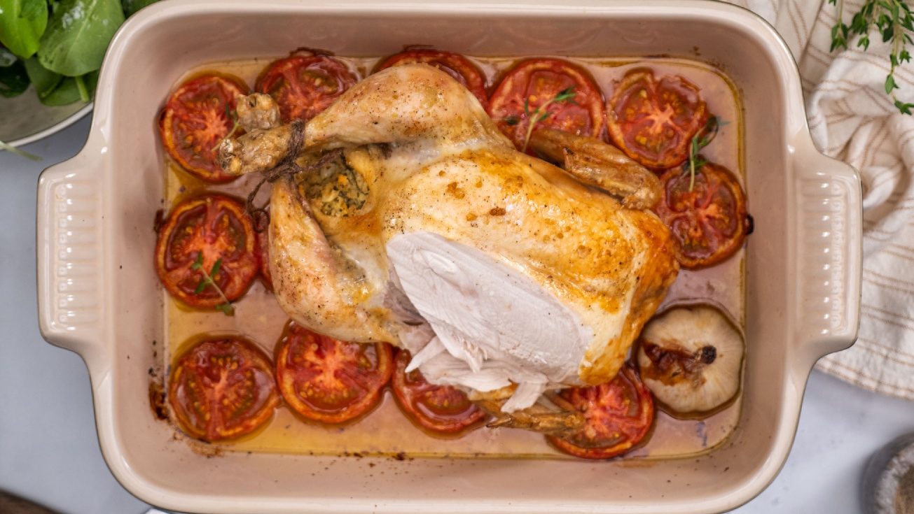 A whole roast chicken set in a deep rectangular baking dish over a bed of sliced tomatoes and a head of garlic.