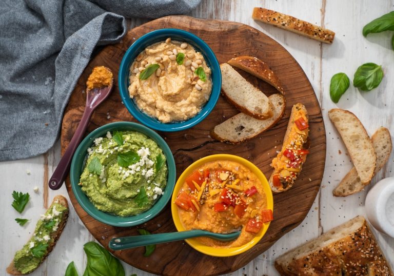 Tree bowles of light brown,green and orange coloured hummus and slices of bread on wooden board