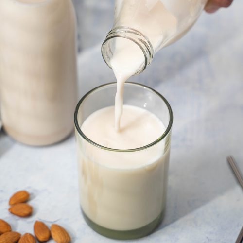 Pouring milk into a glass. A metal straw, almonds, a bottle of milk and green striped cloth