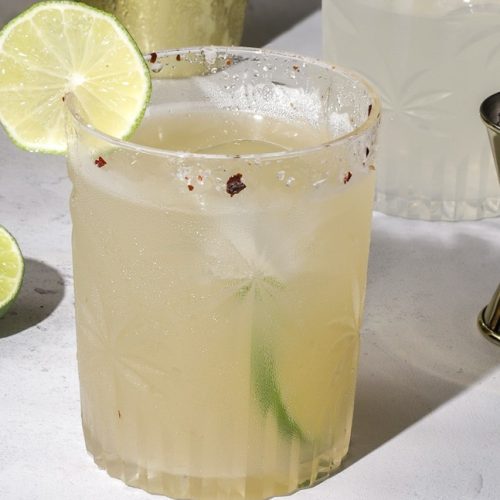 A short glass of margarita with lime slices, and ingredients around it.