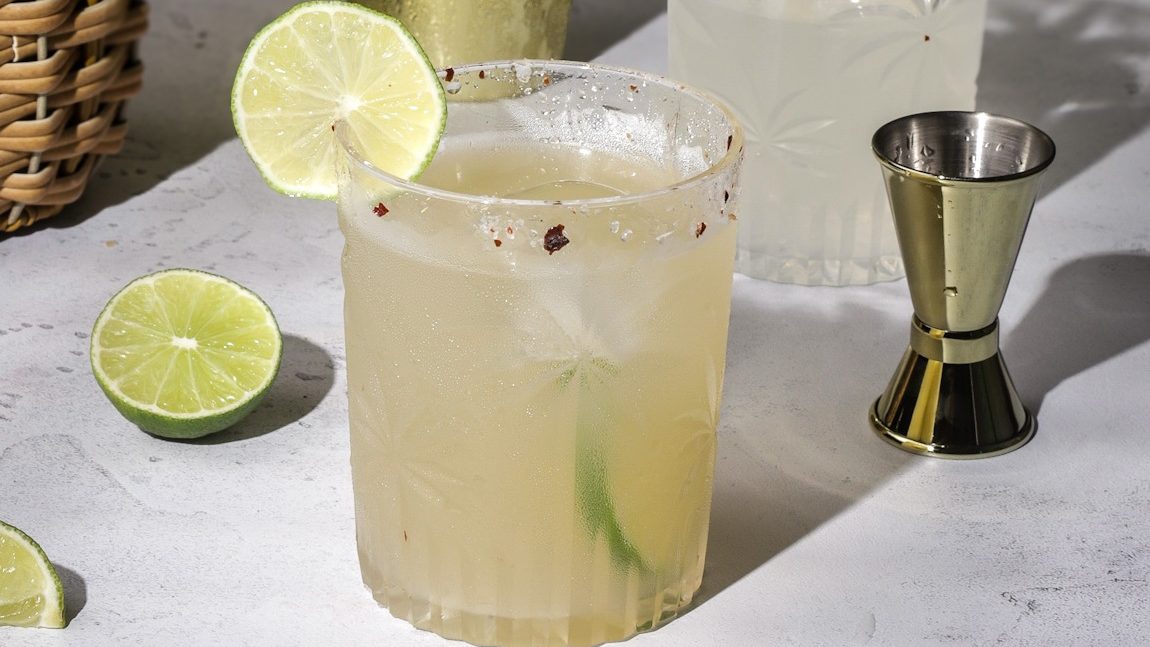A short glass of margarita with lime slices, and ingredients around it.