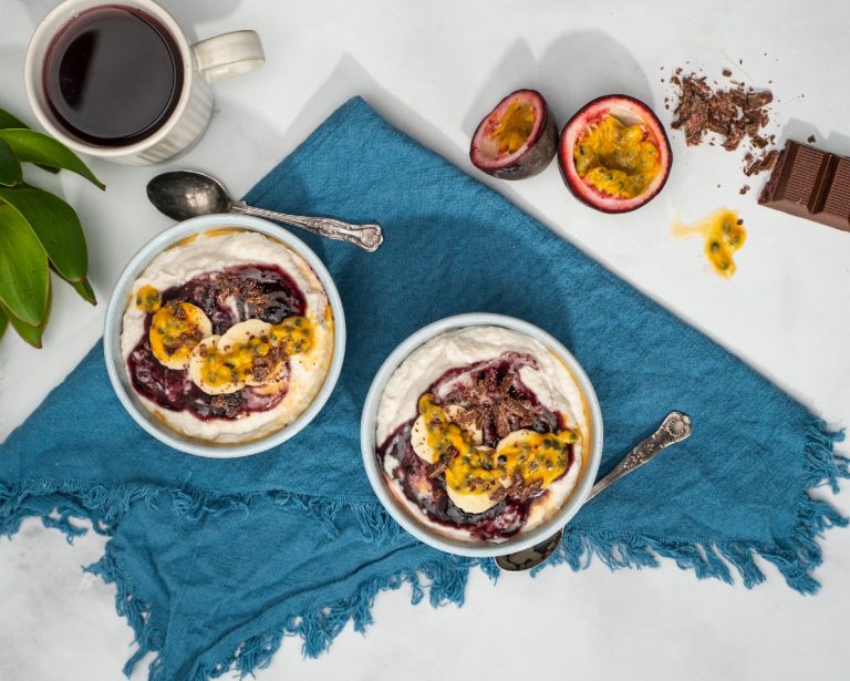 Two bowls of white cream topped with purple jelly and passionfruit pulp on blue mat with a spoon.