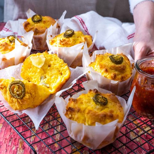 A person showing six yellow muffins with Jalapeno slice on each on wire rack.