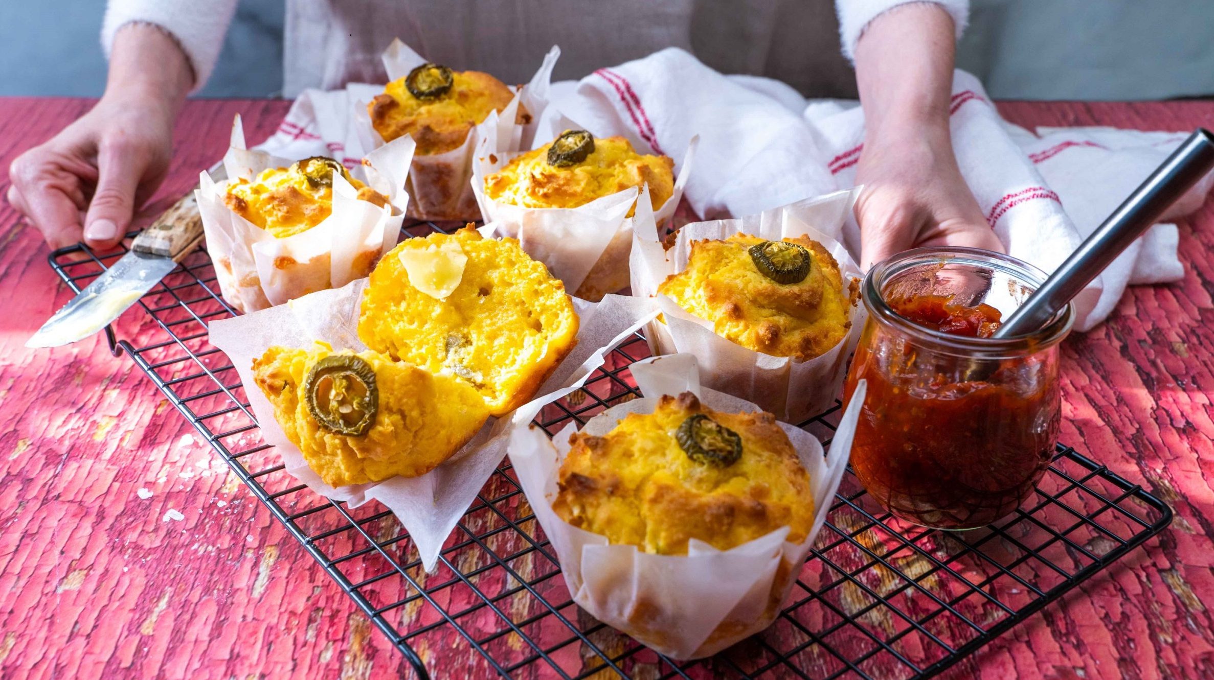 A person showing six yellow muffins with Jalapeno slice on each on wire rack.