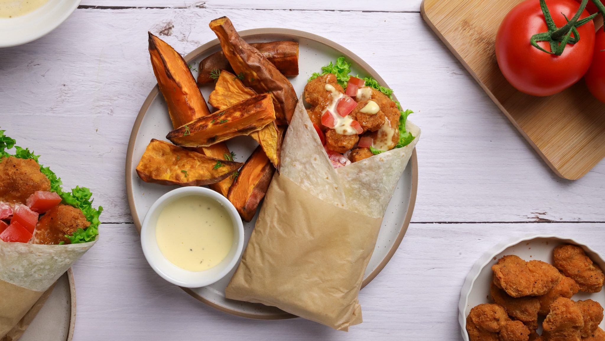 A wrap, showing fried nuggets, lettuce and tomato with mayo on a plate with kumara chips and a pot of creamy sauce.