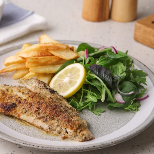 Pan-fried fish fillet with chips, green salad and a lemon slice served on a grey round plate.