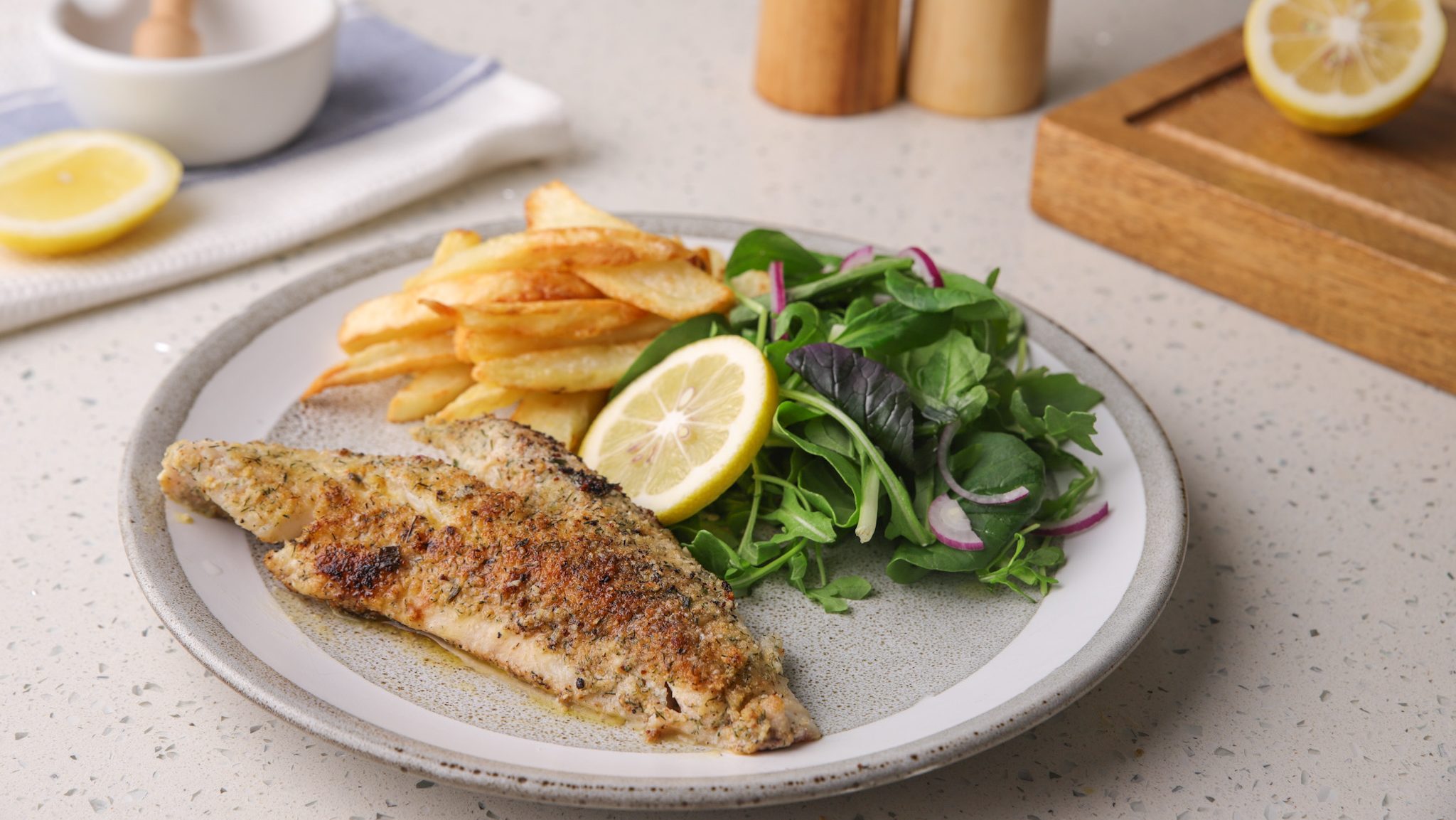 Pan-fried fish fillet with chips, green salad and a lemon slice served on a grey round plate.