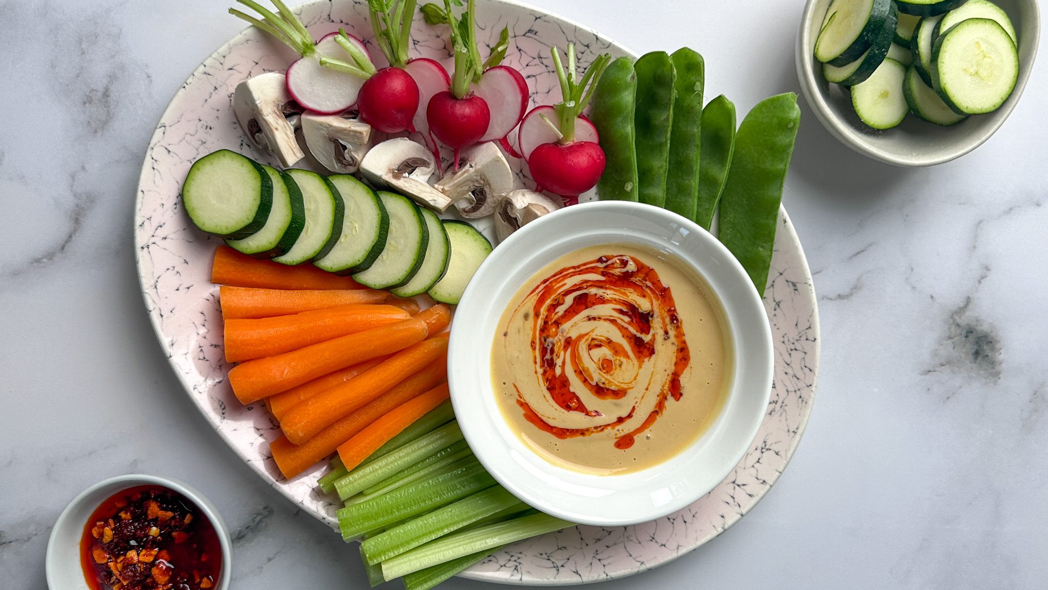 Raw vegetables on a plate with a bowl of light brown sauce with red oil swirl.