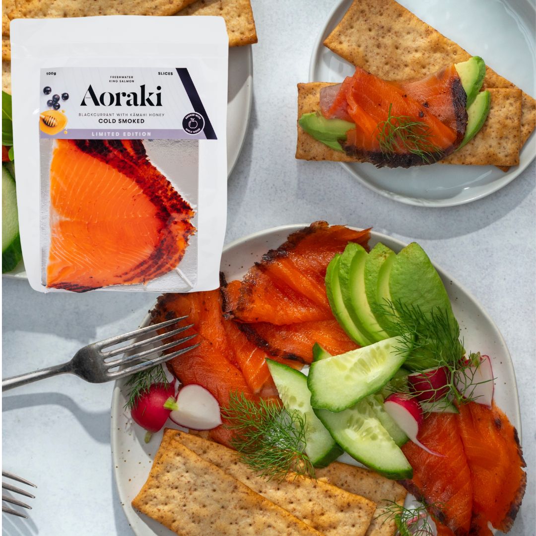 A plate of smoked salmon, crackers, sliced avocado and sliced cucumber with a packet of Aoraki Blackcurrant Cold Smoked Salmon