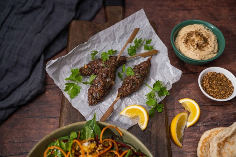 Three meatball skewers, lemon wedges and herb on paper. Small bowls of hummus and dukkah on side.