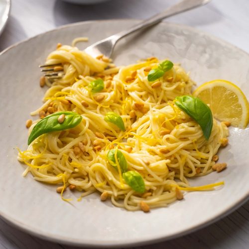 A plate of yellow coloured spaghetti with basil leaves and lemon. A folk full of pasta on it.