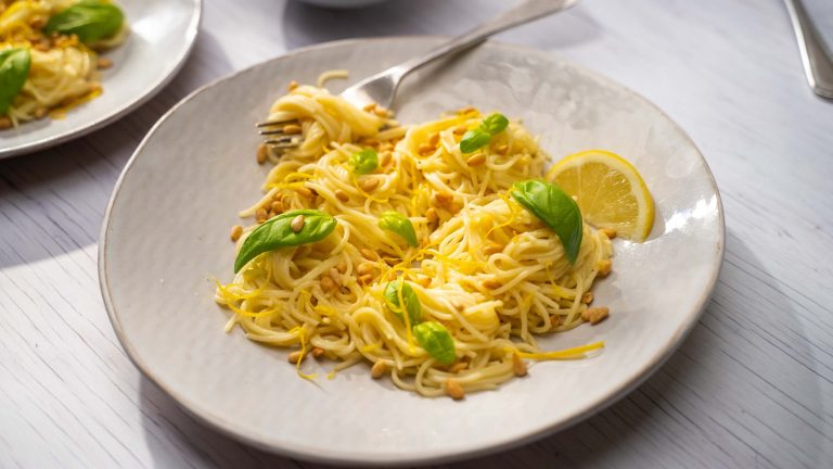 A plate of yellow coloured spaghetti with basil leaves and lemon. A folk full of pasta on it.
