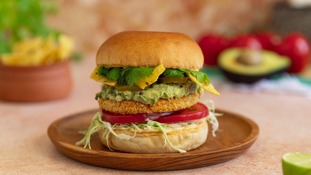 A side view of a crumbed protein burger with tomato, veges mashed avocado on a plate.