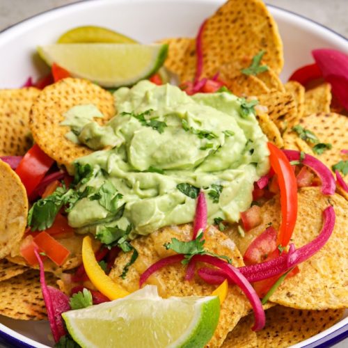 Round corn chips in a bowl with a blob of green cream in the middle, lime wedges and sliced capsicums.