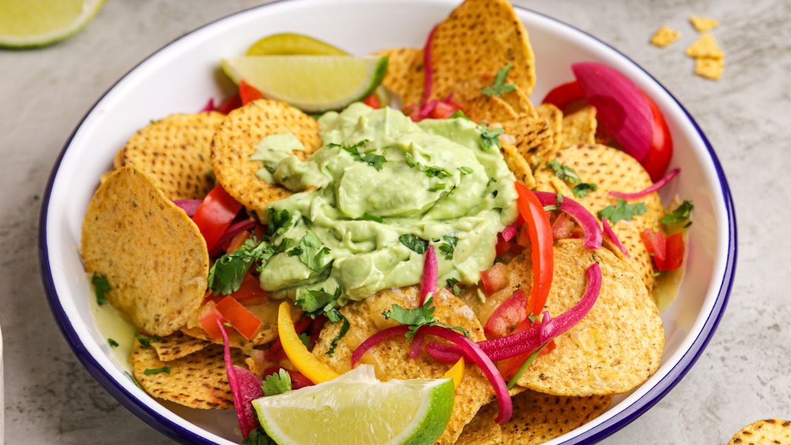 Round corn chips in a bowl with a blob of green cream in the middle, lime wedges and sliced capsicums.