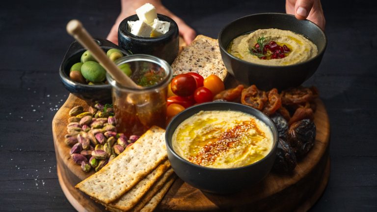 Side view of a grazing platter with two bowls of dips, olives, jar of honey, crackers , nuts and fruits.