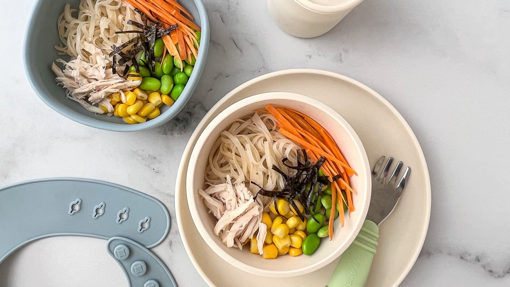Noodles, carrots,chicken, corn, edamame and nori in bowls, with a baby fork and a bib.