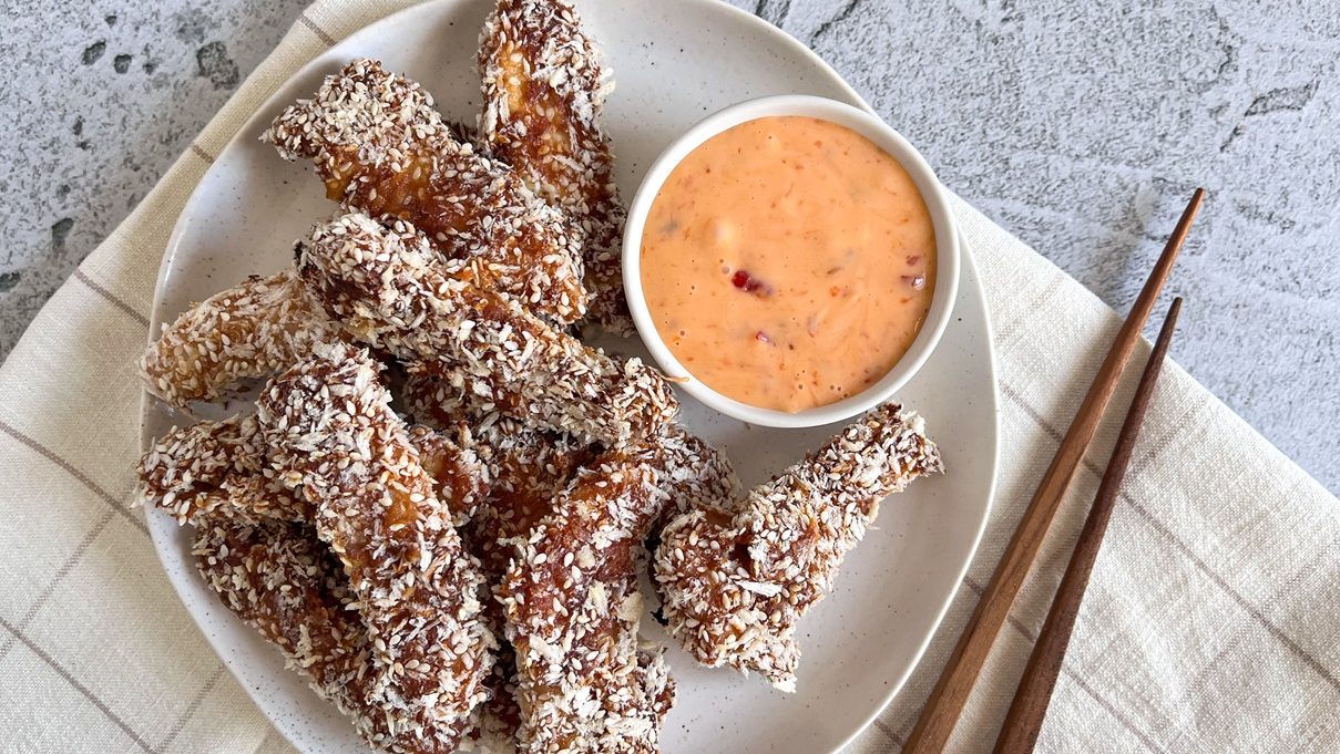 Several crumbed brown pieces of fried food with a bowl of pink sauce on white plate, with a pair of chopsticks.