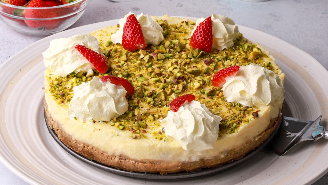 A whole round cheesecake with white cream, strawberries and green nuts on top.