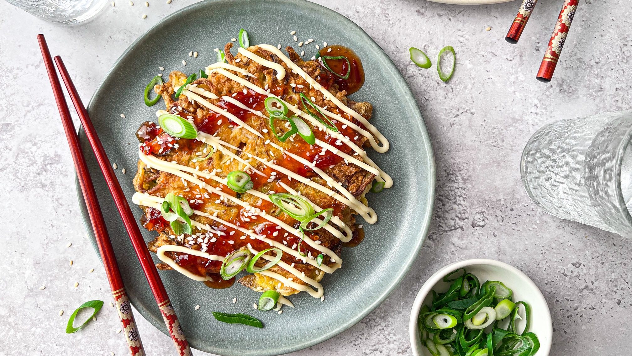 A pancake topped with red sauce, mayo and green onions. With chopsticks.