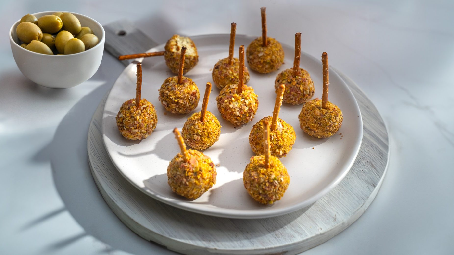 Twelve brown crusted balls with stick on a round white plate with a bowl green olives.