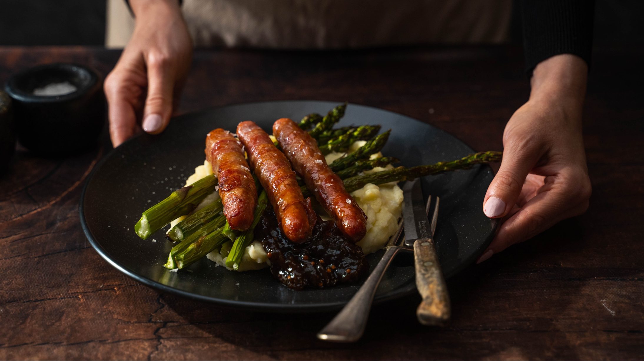 A pair of hands serving a black plate of sausages & mash and green asparagus with a fork and knife.