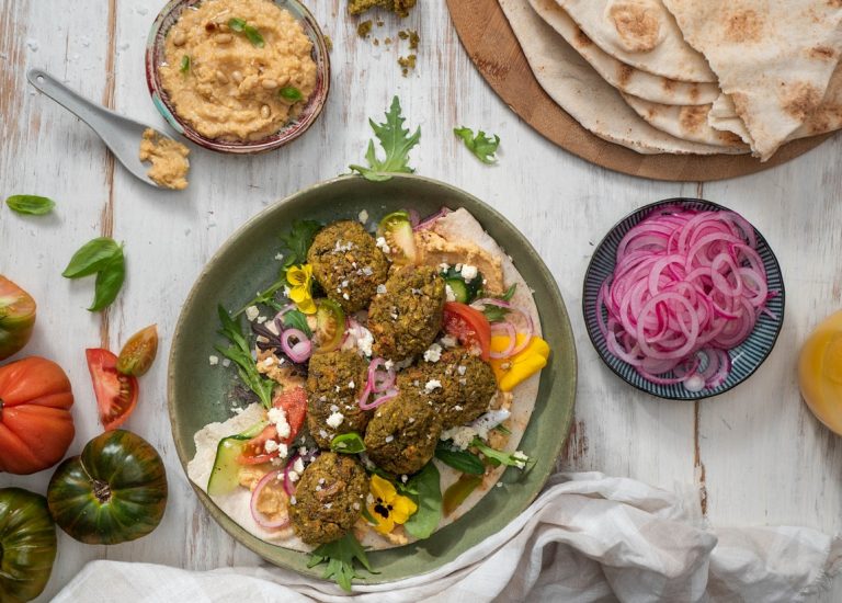 A plate of brown falafel balls on flatbread and red yellow capsicums, a plate of sliced red onions, tomatoes, flatbread around it.