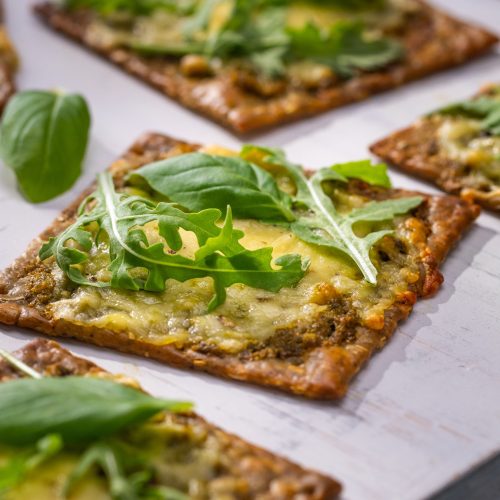 Five square crackers topped with cheese and green rocket leaves on white board.