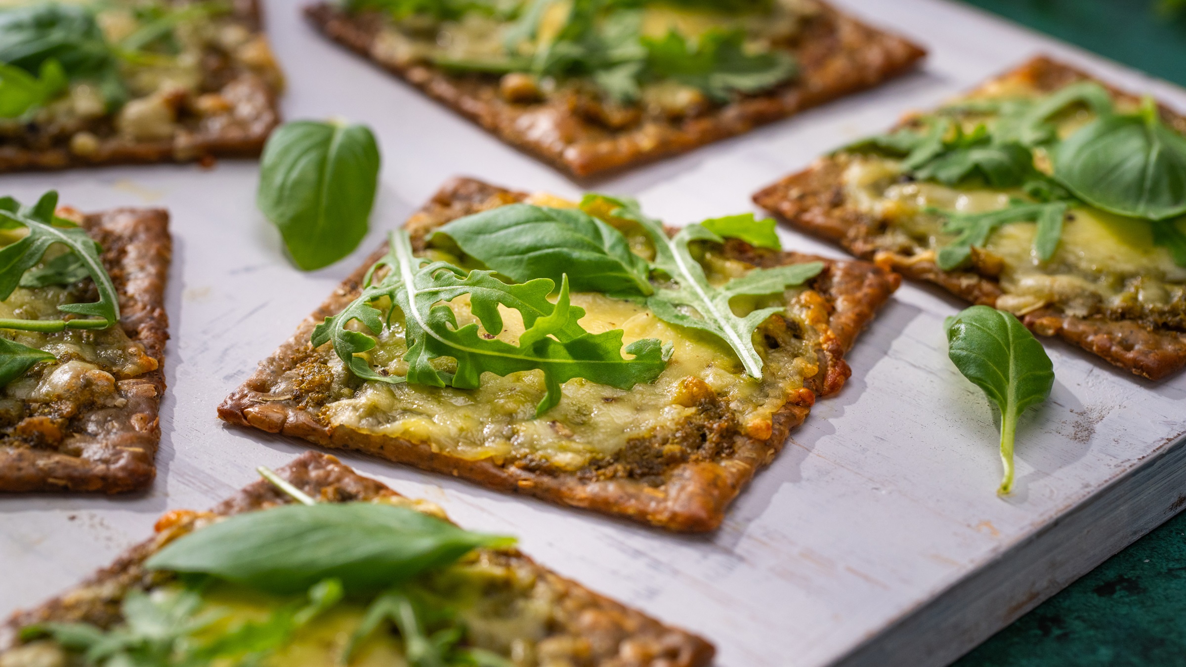 Five square crackers topped with cheese and green rocket leaves on white board.