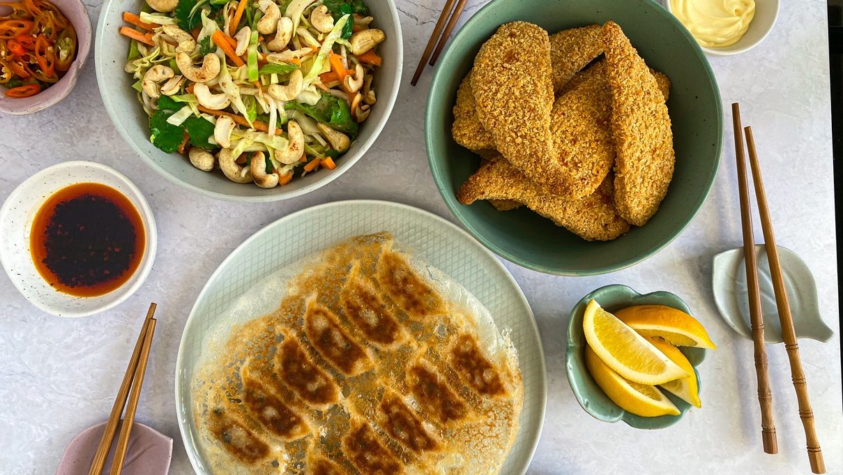 A plate of gyoza, bowl of fried crumbed food, and a bowl of colourful slaw salad, with small bowls of sauces and lemon wedges.