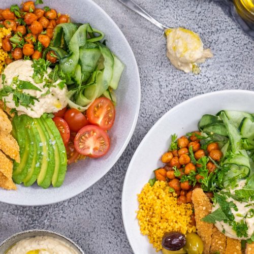 Two buddha bowls on a kitchen top beautifully garnished with avocado, tomatoes, chickpeas, cucumber, olives, couscous and tenders served with hummus