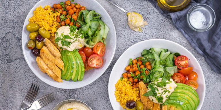 Two buddha bowls on a kitchen top beautifully garnished with avocado, tomatoes, chickpeas, cucumber, olives, couscous and tenders served with hummus