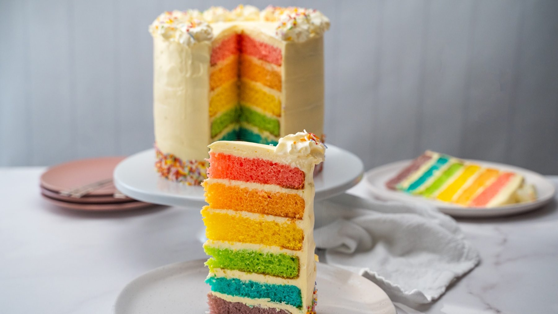 A wedge of tall rainbow coloured sponge layer cake on a plate out of the whole one.