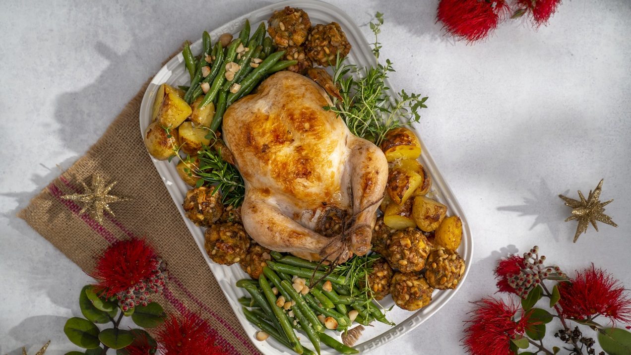 A whole roasted chicken with stuffing balls, green beans and potatoes on a white plate. Pohutakawa flowers around it.