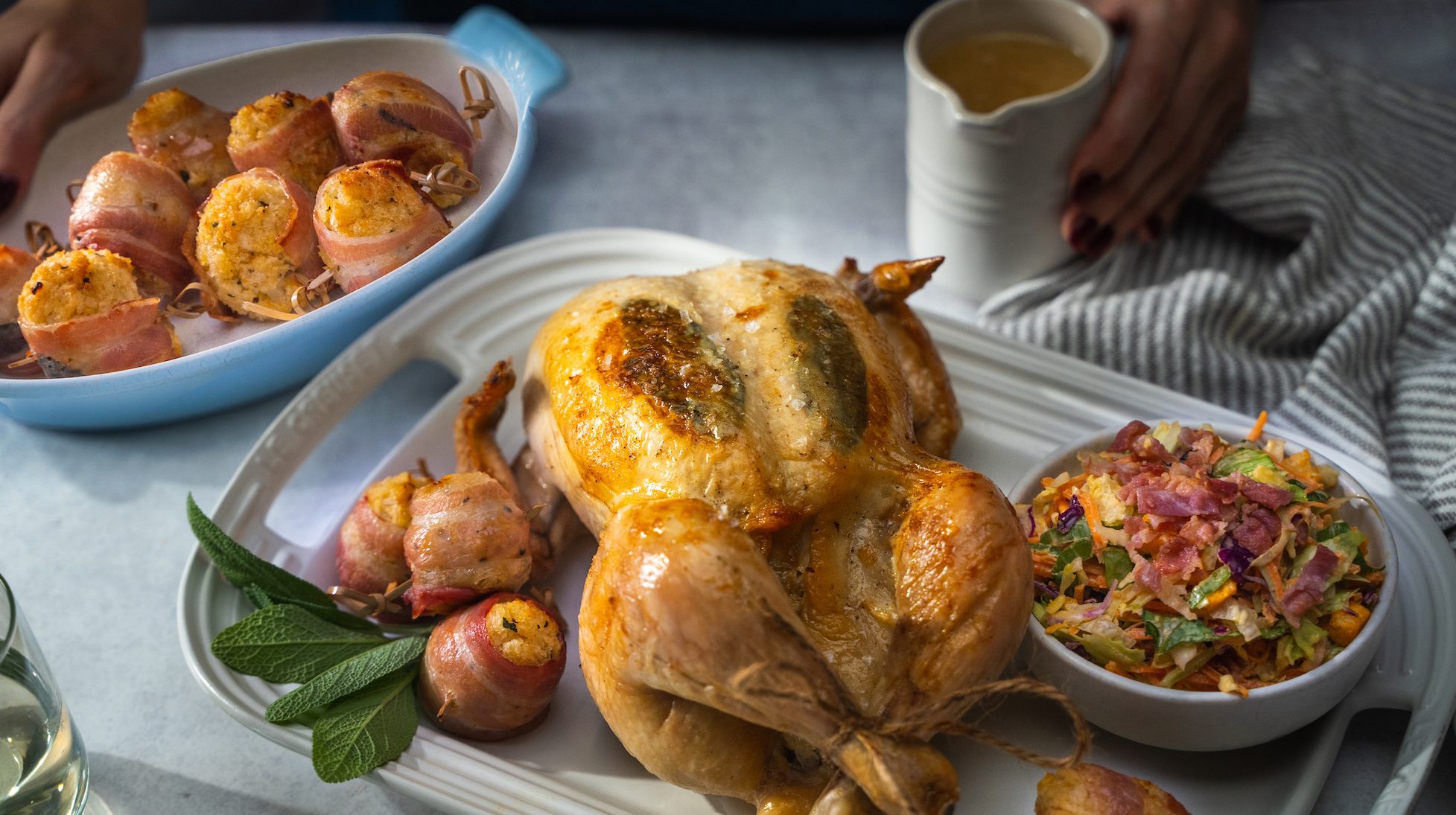 Roast chicken on a tray with bacon-wrapped stuffing balls, a bowl of coleslaw and a jug of gravy