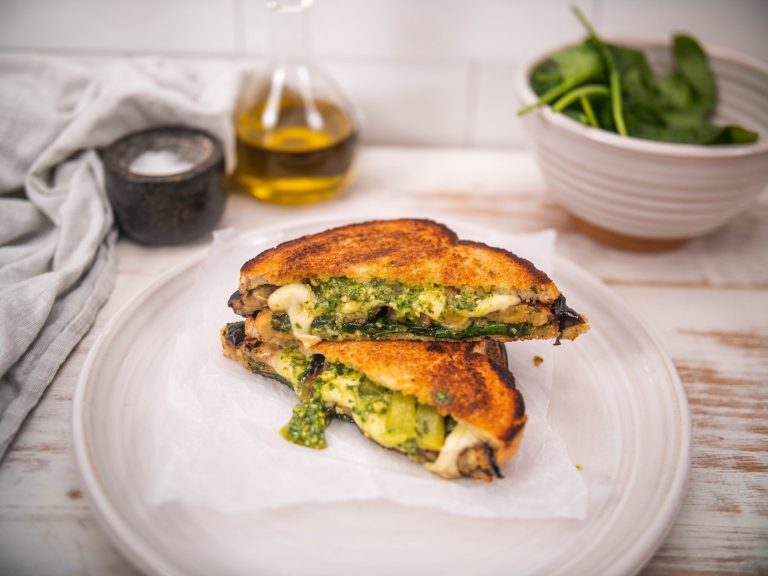 Vegetable toastie with pesto and mozzarella on a white plate with olive oil, salt and greens in the background.