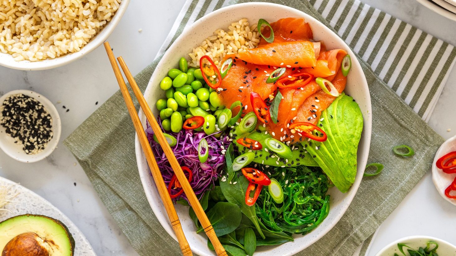 A colourful poke bowl with salmon, avocado, seaweed salad, and edamame. A pair of chopsticks on the bowl.