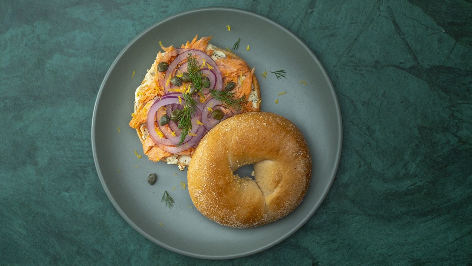 Bagel with salmon flake, sliced red onion, herb and capers on green plate.