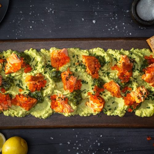 Green avocado pasted on wooden board, dotted with salmon flesh, roes and herbs. Lemon halves and rectangler crackers on side.