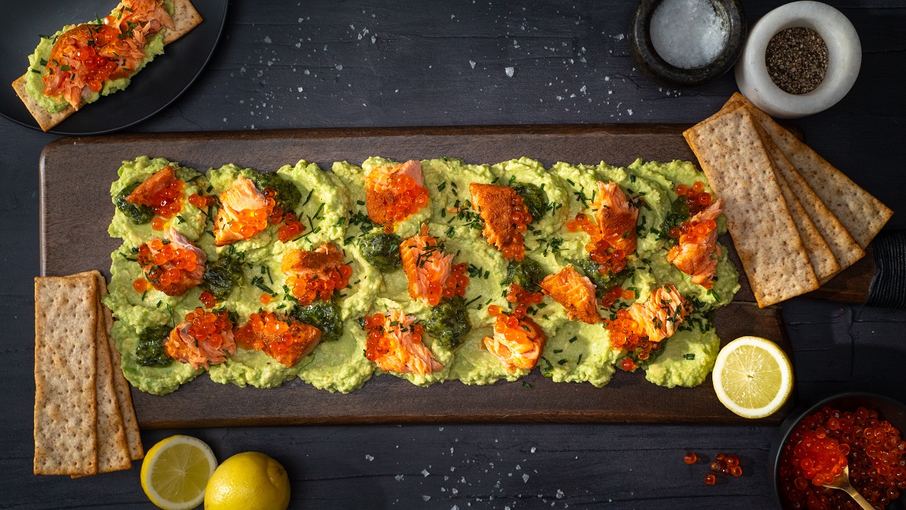 Green avocado pasted on wooden board, dotted with salmon flesh, roes and herbs. Lemon halves and rectangler crackers on side.