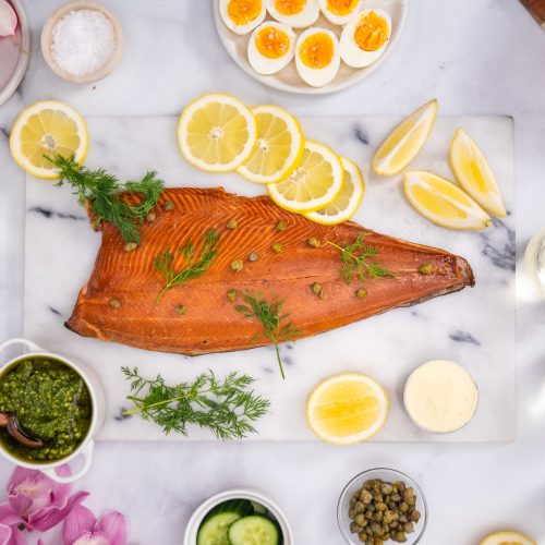 Smoked salmon side on a marble platter served with fresh dill, lemon slices, capers and a pottle of rich pesto and pine nut sauce.
