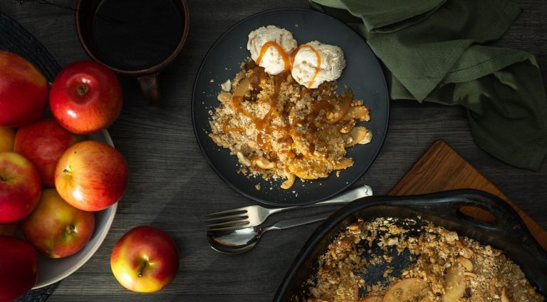 Several red apples in a bowl on left, brown crumble with two scoops of ice cream drizzled with brown sauce on blue plate in middle and black baking dish with crumble on right.