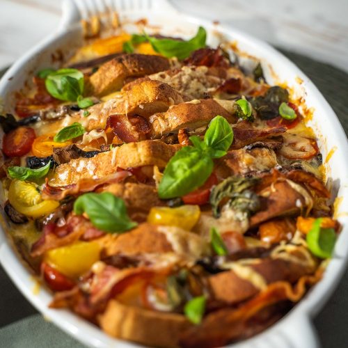 A white oven dish full of bread, bacon, tomato, mushroom bake topped with basil leaves.