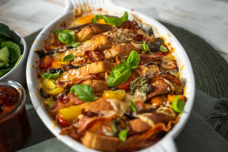 A white oven dish full of bread, bacon, tomato, mushroom bake topped with basil leaves.