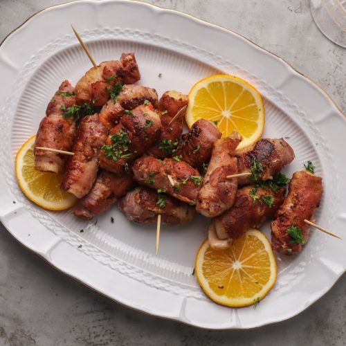 Bacon wrapped foods with toothpicks on a white oval plate with lemon slices and herb.
