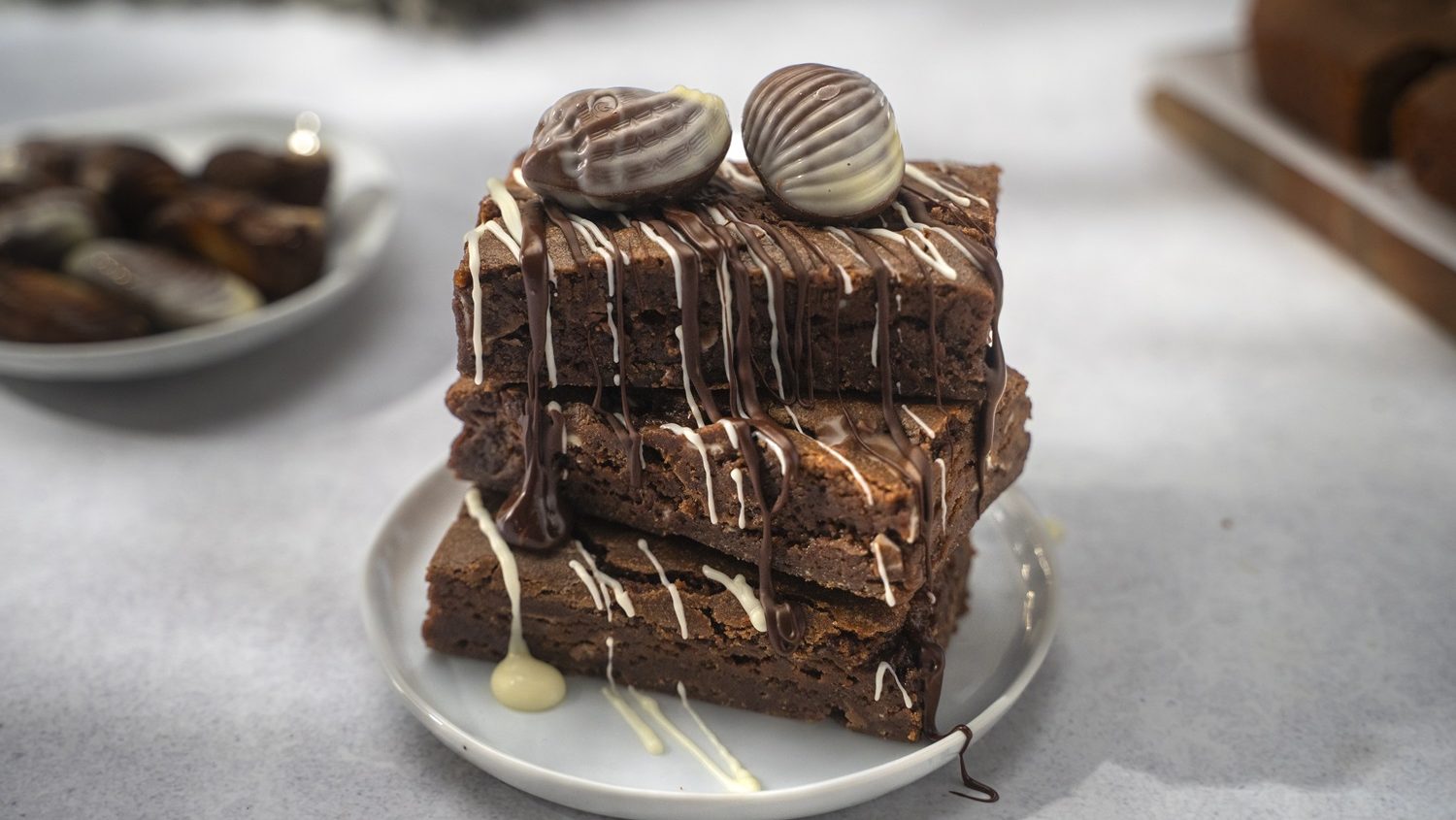 Three pieces of chocolate brownies stack on a plate, topped with chocolate drizzle and two shells chocolates.