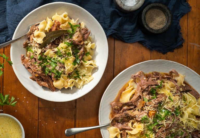 two bowls of pasta topped with lamb and herb. Two small pots of salt and pepper in the back. On wooden board.