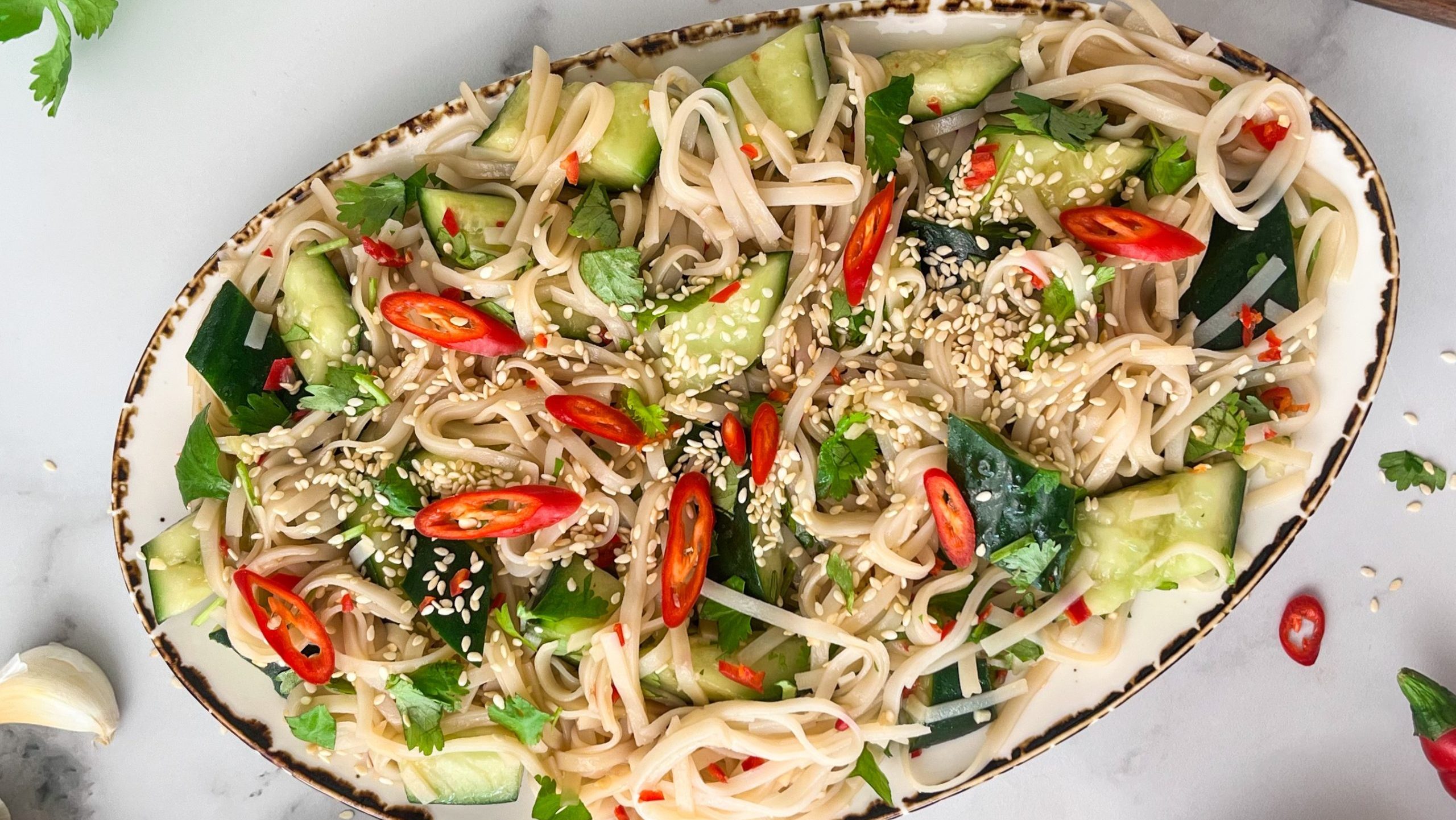An oval plate full of noodles topped with cucumber pieces, green herbs, red chilli and sesame seeds.